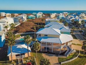 A coastal residential area with houses featuring blue roofs and white exteriors, a central house with a balcony, and a clear view of the ocean. - Shared Pool, Boardwalk to Beach and Golf Cart Included (Port Aransas)