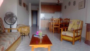 Uma sala de estar com sofá, uma mesa de madeira com um vaso de flores, uma poltrona de vime e um espaço de jantar com cadeiras e mesa de madeira.
