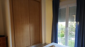 Quarto com duas camas, um guarda-roupa, uma janela com persianas e uma varanda com vista.