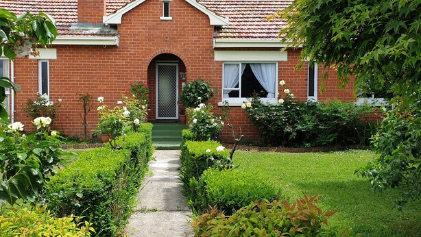 A brick house with a green door, white windows, and a garden with various plants and flowers. - A cozy art deco farmhouse within walking distances to all amenities in town. (Te Awamutu)
