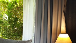 A neatly made bed with a patterned bedspread and pillows, a bedside lamp, and a view of greenery through sheer curtains. - A cozy art deco farmhouse within walking distances to all amenities in town. (Te Awamutu)
