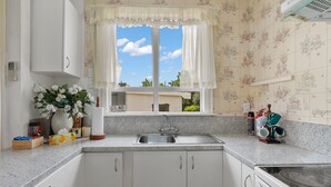 A kitchen with white cabinets, a stainless steel sink, a window with floral curtains, and a view of the outside. - A cozy art deco farmhouse within walking distances to all amenities in town. (Te Awamutu)