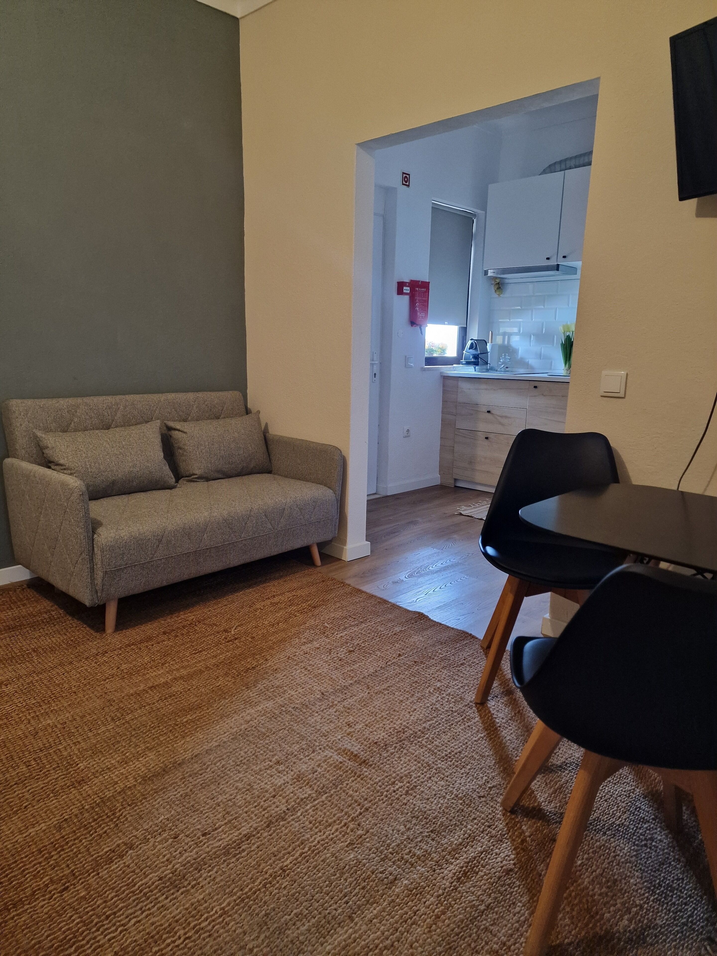 Una sala de estar con un sofá, una mesa de comedor con sillas, una zona de cocina visible a través de una puerta abierta y una alfombra estampada en el suelo.
