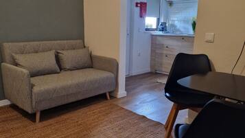 Una sala de estar con un sofá, una mesa de comedor con sillas, una zona de cocina visible a través de una puerta abierta y una alfombra estampada en el suelo.