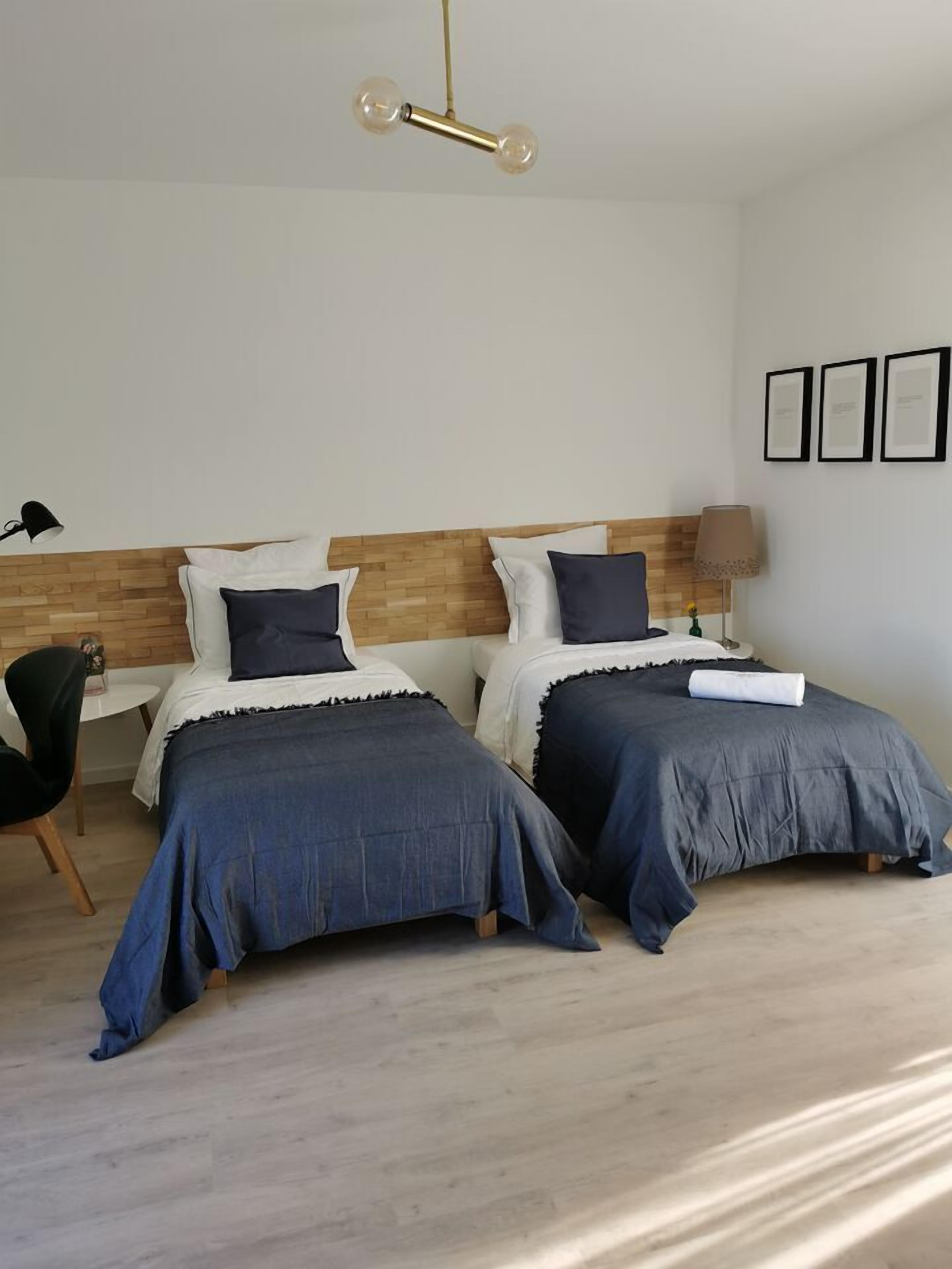 Ein modernes Schlafzimmer mit zwei Betten, einem hölzernen Kopfende, einer runden Deckenleuchte, einem Sessel und einem kleinen Tisch.