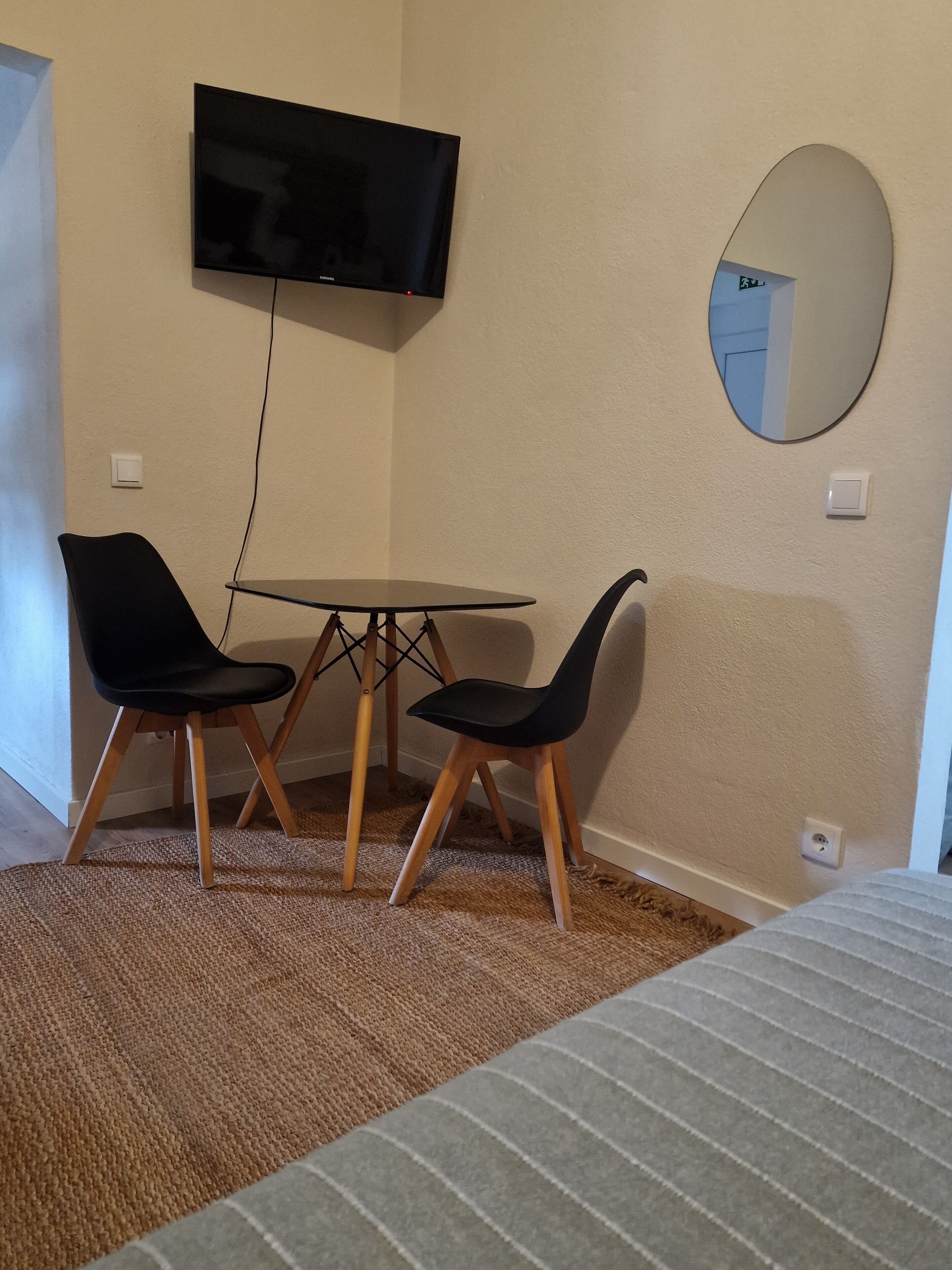 A room with a bed, a TV mounted on the wall, a round mirror, and two chairs around a small table.