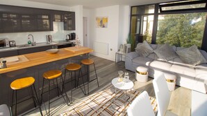 Een moderne keuken met een centraal eiland, barkrukken, een bank en uitzicht op groen door grote ramen.