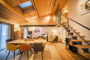 Amplia sala de estar con techo de madera, una mesa de comedor con sillas, una escalera y vista del salón a través de una puerta abierta.