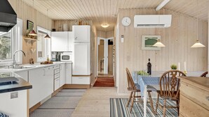 A modern kitchen with white cabinets, a microwave, and a refrigerator. There is a dining area with a table and chairs, and a clock on the wall. - 3 bedroom stunning home in Juelsminde (Juelsminde)