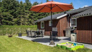 A wooden house with a patio, outdoor grill, and a play area with sand and toys. - 3 bedroom stunning home in Juelsminde (Juelsminde)