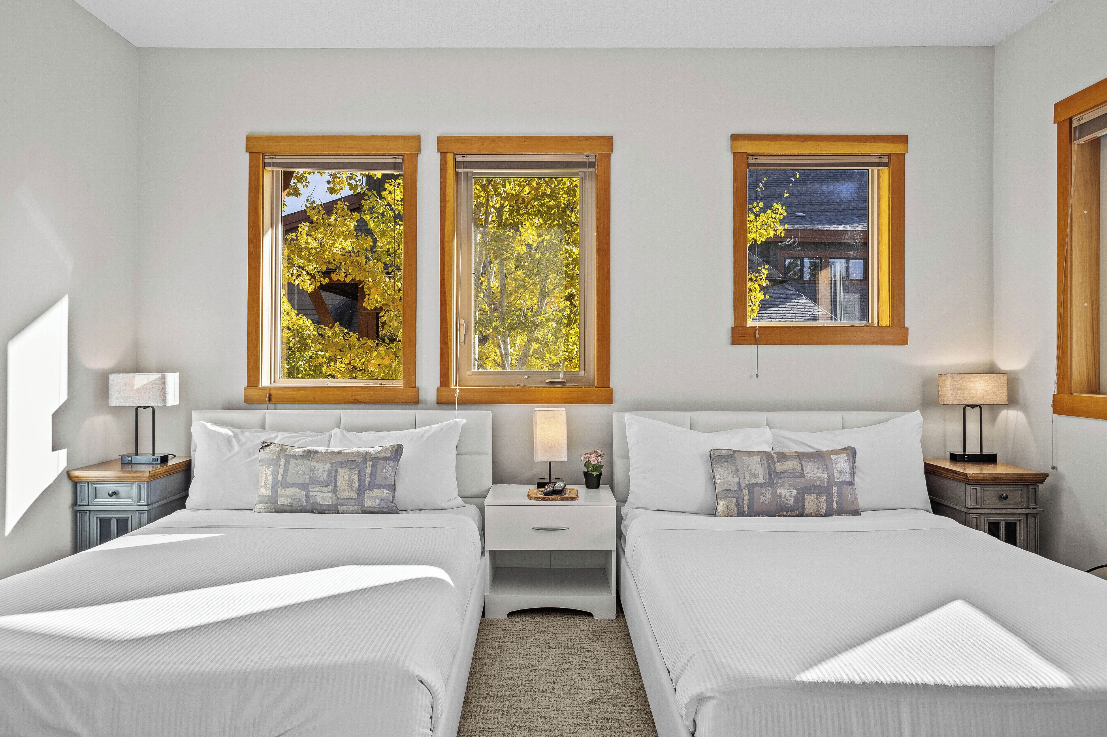 Une chambre d’hôtel avec deux lits, une table de chevet avec une lampe et trois fenêtres donnant sur des arbres et un immeuble.