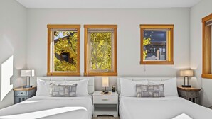 Une chambre d’hôtel avec deux lits, une table de chevet avec une lampe et trois fenêtres donnant sur des arbres et un immeuble.