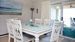A dining area with a glass-top table, white chairs, a ceiling fan, a painting of a beach, and a view through a glass door to an outdoor deck. - Stylish, cozy 2 bedroom luxury oceanfront condo with free WiFi, an outdoor pool, and a stunning ocean view from a covered balcony and only steps from the beach in midtown! (Ocean City)