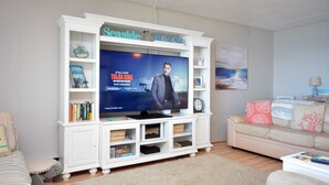 A living room with a television on a white entertainment unit, a beige sofa, and a small table with books. - Stylish, cozy 2 bedroom luxury oceanfront condo with free WiFi, an outdoor pool, and a stunning ocean view from a covered balcony and only steps from the beach in midtown! (Ocean City)