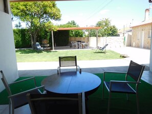 Un patio con una mesa redonda y sillas, un área de estar techada, y vista a un jardín con césped y árboles.