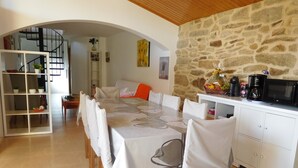 A dining area with a long table, white chairs, and a stone wall. There is a staircase in the background, and a kitchen area with a microwave and coffee maker. - Casa con Jardin en el Salnes (Meaño)
