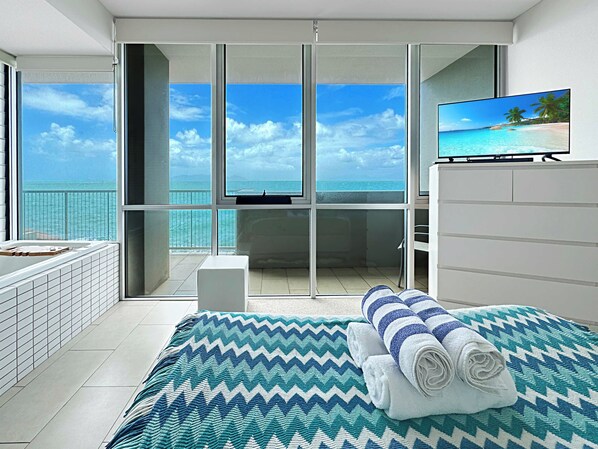 A hotel room with a large balcony, a bed with a patterned bedspread, a TV, and a view of the sea.