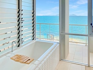 A modern bathroom with a bathtub, a wooden tray, and a view of the sea through a balcony.