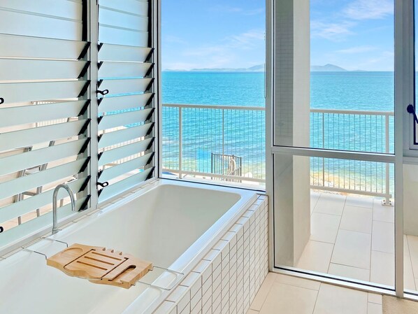 A modern bathroom with a bathtub, a wooden tray, and a view of the sea through a balcony.