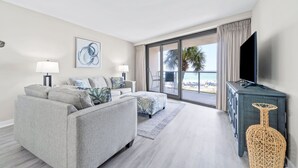 Ein modernes Wohnzimmer mit einer Couch, zwei Beistelltischen, einem Flachbildfernseher und einem großen Fenster mit Blick auf den Strand.