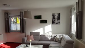 Un saló modern amb un sofà gris, una taula de café blanca i una làmpada de peu. Hi ha dues finestres amb cortines, un radiador i un quadre penjat a la paret.