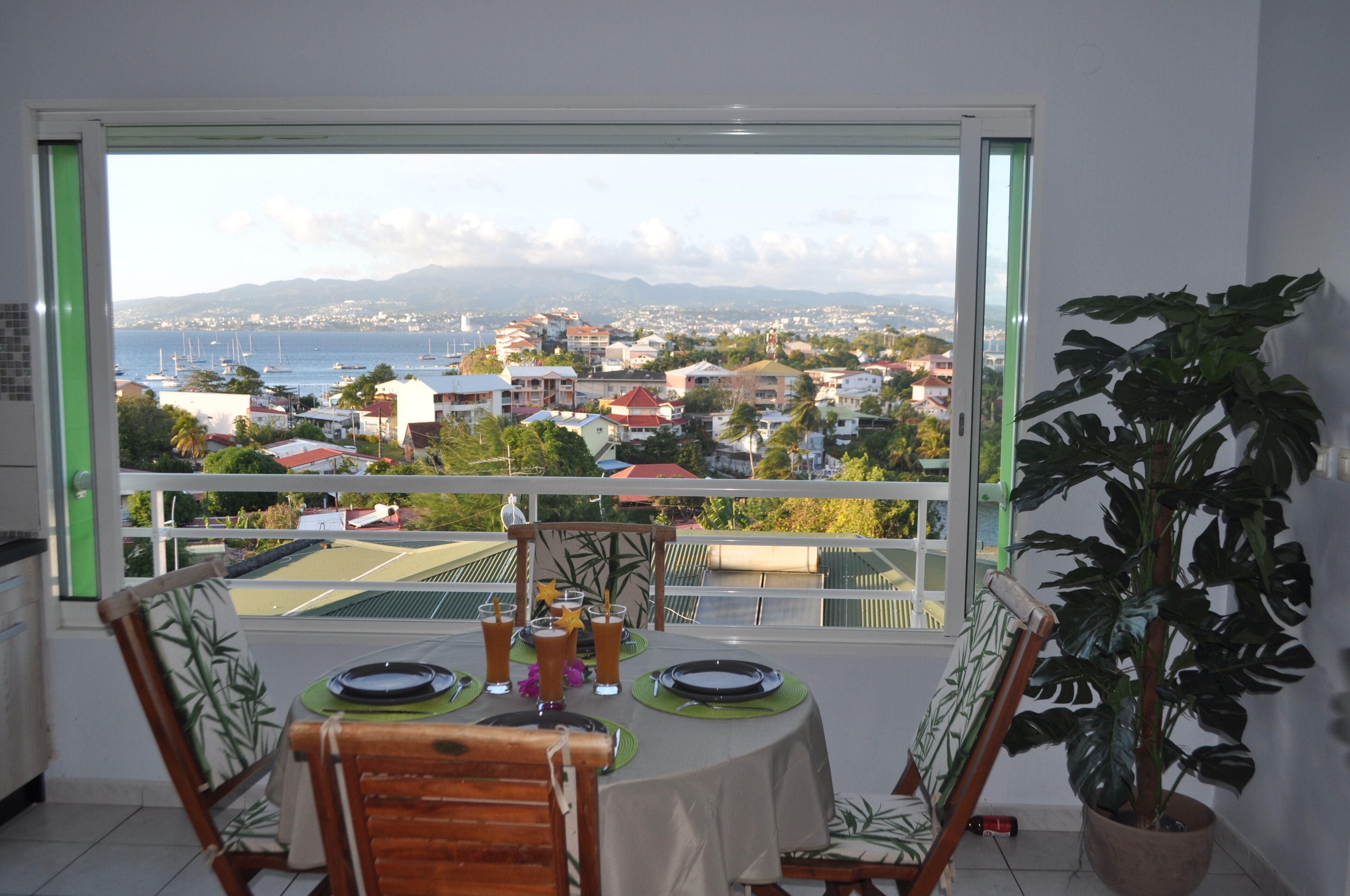 A dining area with a table set for two, a potted plant, and a view of a coastal town through a window.