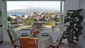 A dining area with a table set for two, a potted plant, and a view of a coastal town through a window. - L'R2 LA VILLA MELISSA T2 APARTMENT WITH POOL 150 M FROM THE BEACH. (Les Trois-Îlets)