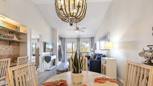 A dining area with a round table, chairs, and a chandelier. There is a plant in the center of the table, and the room has a ceiling fan.
