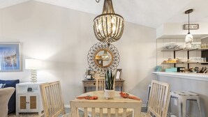 A dining area with a wooden table, chairs, a chandelier, and a kitchen area with shelves and appliances.