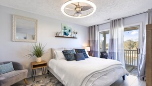 A bedroom with a large bed, a side table with a plant, a mirror, and a ceiling fan.