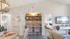 A modern living room with a dining area, a kitchen with a tiled backsplash, and a television mounted on the wall.