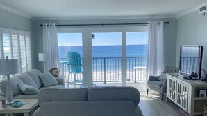 Una sala de estar con un sofá gris, una mesita, un televisor y vistas a la playa a través de amplios ventanales.