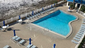 Un área de piscina con sillones de playa y sombrillas, una playa al fondo y un edificio con techo azul.