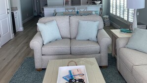 Una sala de estar con un sofá color beige, una mesa de centro con una bandeja, y una cocina al fondo.