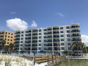 A multi-story hotel with balconies, a sandy beach area with umbrellas, and a white fence.
