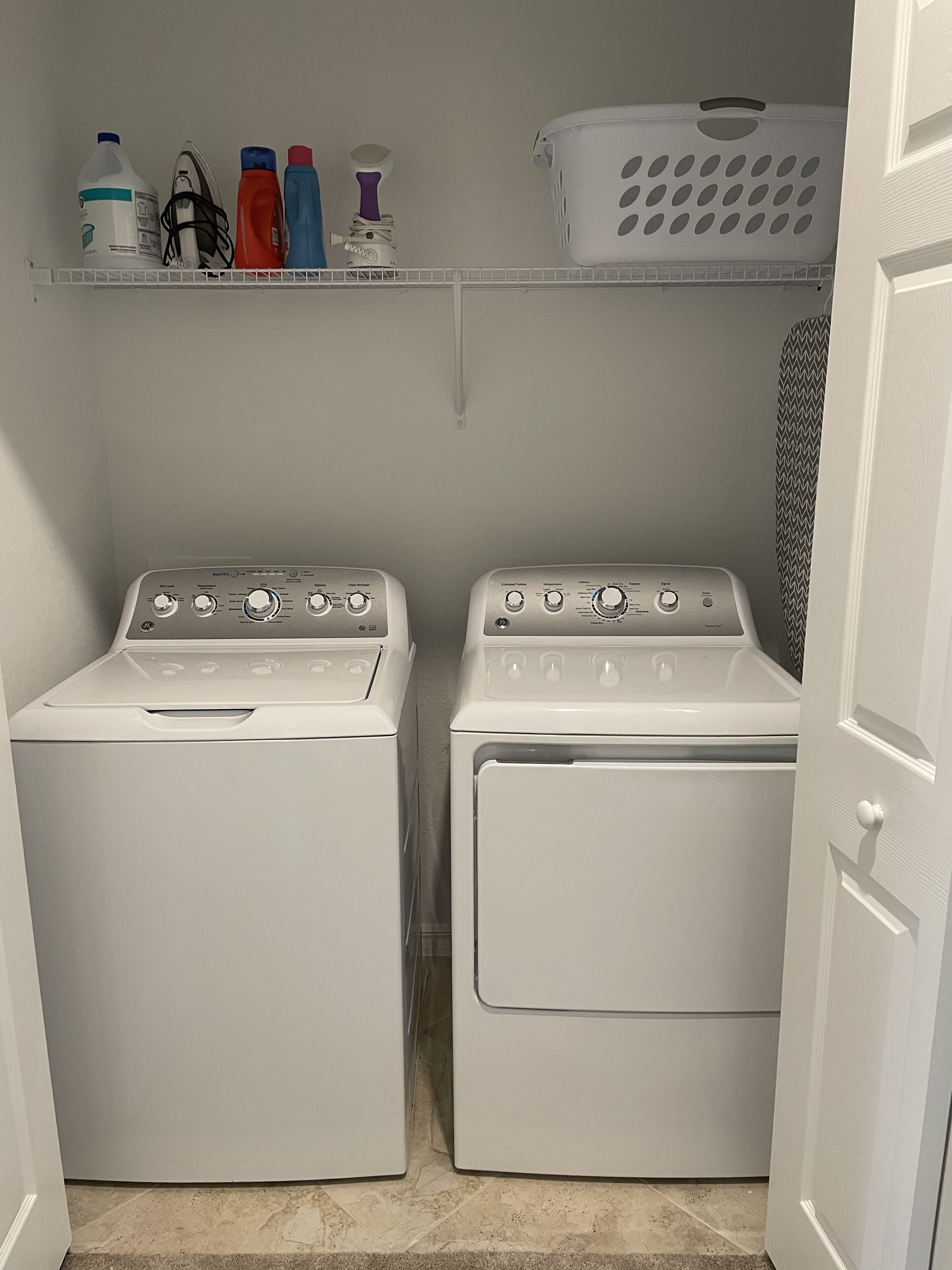 A laundry room with two white front-loading washing machines, a shelf above with various laundry supplies, and a white laundry basket.