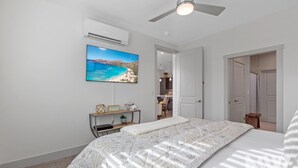 Ein Schlafzimmer mit einem Bett, einer Wand-Klimaanlage, einem Deckenventilator, einem Strandbild und Blick ins angrenzende Badezimmer.