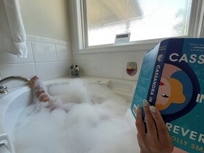 Una vasca da bagno piena di bolle, una finestra con vista e un libro intitolato 'CASSANDRA IN REVERIE' di Holly Smaele.
