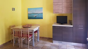 Un coin repas avec une table et des chaises, un tableau représentant des bateaux sur l’eau, un téléviseur posé sur un meuble et un réfrigérateur.