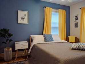 Ein Schlafzimmer mit einem Bett, einem Nachttisch, einem Fenster mit gelben Vorhängen und einem Wandbild.