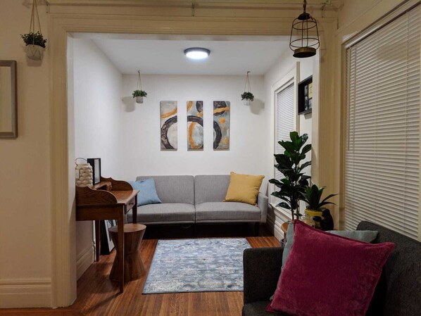 Una sala de estar con un sofá gris, una mesa de centro de madera y un tapete azul. Hay tres cuadros abstractos en la pared y una planta en maceta.