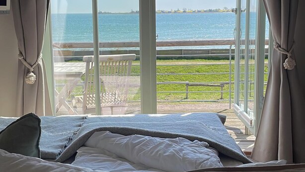 A bedroom with a large window overlooking a body of water and a distant shoreline. - Strandhaus-Fehmarn, port side, sea view and beach on the doorstep (Fehmarn)