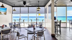 Ein Restaurant mit Tischen und Stühlen, großen Fenstern mit Meerblick, Deckenventilatoren und Pendelleuchten.