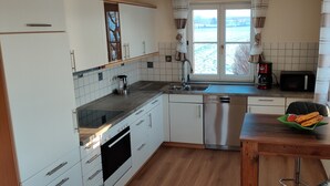 A modern kitchen with white cabinets, stainless steel appliances, a window with a view, and a wooden table with fruit. - 🌞FeWo Berger, quiet location on the outskirts of Frauenzell, near Center Parcs in the Allgäu (Altusried - Frauenzell)