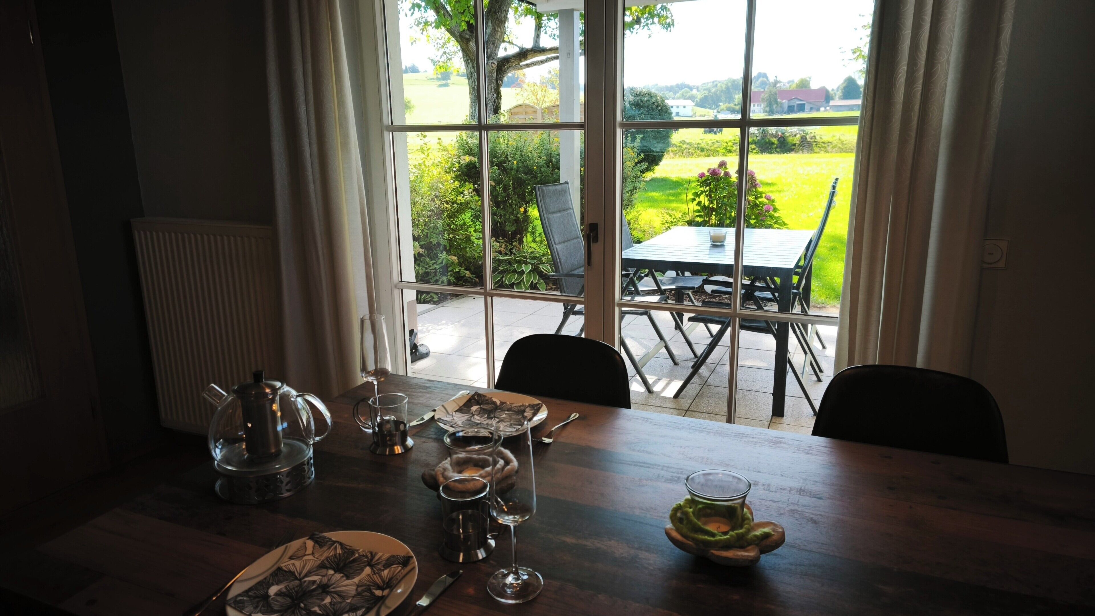 A dining area with a table set for two, a view of a garden through large windows, and a patio with outdoor seating.