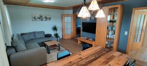 Un salon moderne avec un canapé gris, une table basse en bois et une table à manger avec des chaises. Un téléviseur est fixé au mur, il y a des meubles en bois et une plante en pot.