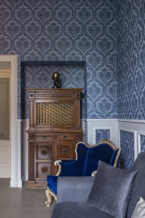 Una stanza con carta da parati decorata, un armadietto in legno con cassetti, una poltrona rivestita di blu e un divano grigio.