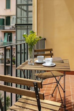 Un tavolo di legno con due tazze e piattini bianchi, un vaso con piante verdi e una panca su un balcone con pavimento piastrellato.