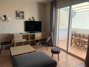 Une chambre moderne dotée d’un téléviseur à écran plat, d’un bureau en bois, de deux fauteuils en osier, d’un canapé gris et d’une porte-fenêtre coulissante donnant sur un espace extérieur aménagé avec du mobilier en bois.