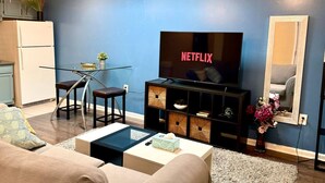 A living room with a TV displaying 'NETFLIX', a glass dining table, a sofa, and a kitchen area in the background. - Cozy 1BD Apt @Magic Garden in Center City (Philadelphia)